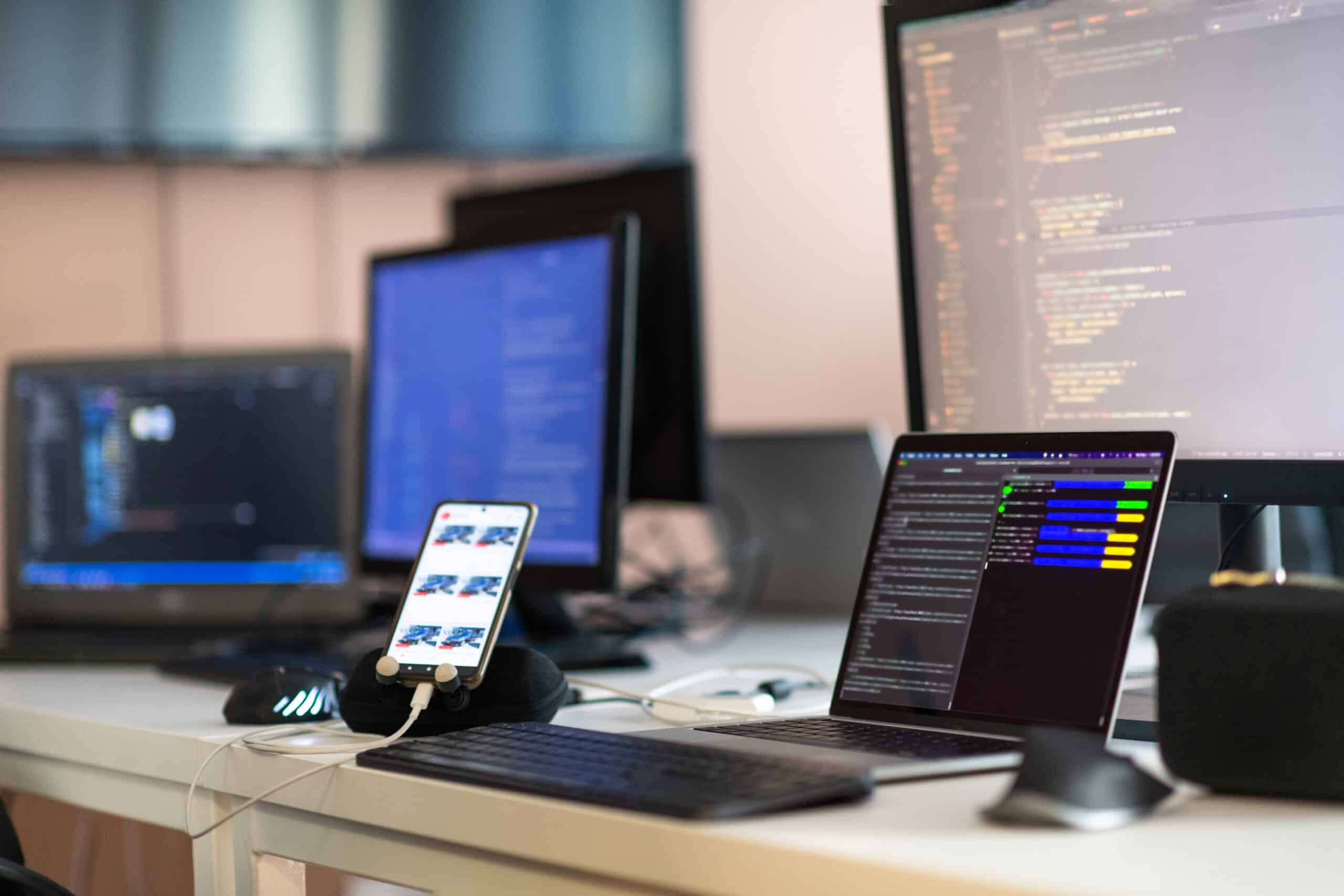 Ein Arbeitsplatz mit einem Laptop, einem Smartphone und einem Monitor, der Code anzeigt. Der Schreibtisch ist mit verschiedenen technischen Geräten ausgestattet.